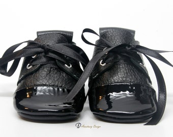 May include: A pair of black leather baby shoes with black satin laces. The shoes have a patent leather toe cap.