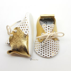 Puede incluir: Un par de zapatos de beb&eacute; de cuero blanco con lunares dorados y detalles de cuero dorado. Los zapatos tienen cordones y est&aacute;n atados.