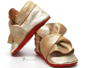 May include: Gold leather baby shoes with a red sole and a large bow on the front. The shoes are made of soft, supple leather and are perfect for a special occasion.