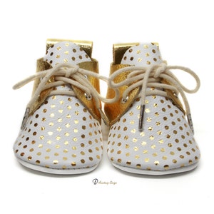 Puede incluir: Un par de zapatos de beb&eacute; de cuero blanco con lunares dorados y detalles de cuero dorado. Los zapatos tienen cordones y una suela blanca.