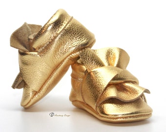 May include: A pair of gold leather baby shoes with a ruffled bow detail. The shoes are made of soft, supple leather and have a comfortable fit.