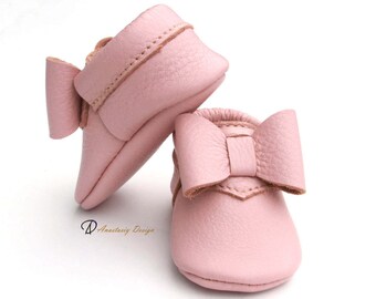 May include: A pair of pink leather baby shoes with a bow on the front. The shoes are soft and flexible, ideal for little feet.