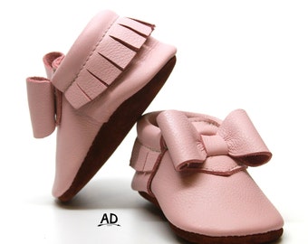 May include: A pair of pink leather baby moccasins with a bow on the front and fringe detail.