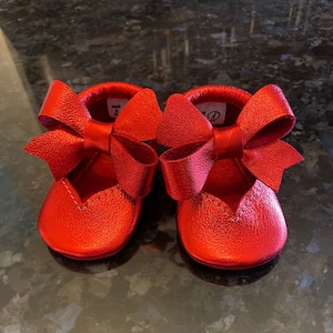 K&ouml;nnte beinhalten: Ein Paar rote Lederschuhe f&uuml;r Babys mit einer gro&szlig;en Schleife an jedem Schuh.