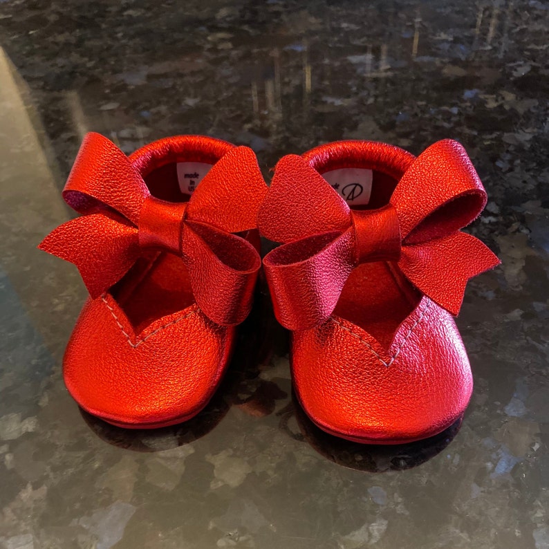 K&ouml;nnte beinhalten: Ein Paar rote Lederschuhe f&uuml;r Babys mit einer gro&szlig;en Schleife an jedem Schuh.