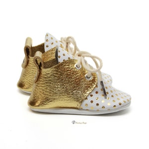 Puede incluir: Un par de zapatos de beb&eacute; de cuero blanco con lunares dorados y detalles en cuero dorado. Los zapatos tienen cordones y una punta redonda.