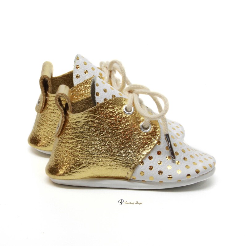 Puede incluir: Un par de zapatos de beb&eacute; de cuero blanco con lunares dorados y detalles en cuero dorado. Los zapatos tienen cordones y una punta redonda.