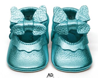 May include: A pair of teal blue leather baby shoes with a bow on the top. The shoes have a scalloped edge and are made of soft leather.