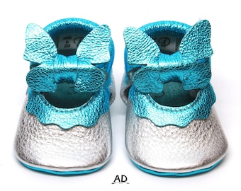May include: A pair of baby shoes with a silver and turquoise metallic finish. The shoes have a bow on the front and are made in the USA.
