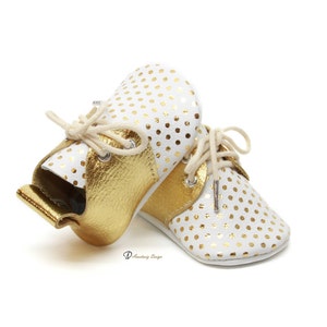Puede incluir: Un par de zapatos de beb&eacute; de cuero blanco con lunares dorados y detalles de cuero dorado. Los zapatos tienen cordones y una punta redonda.