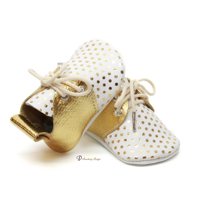 Puede incluir: Un par de zapatos de beb&eacute; de cuero blanco con lunares dorados y detalles de cuero dorado. Los zapatos tienen cordones y una punta redonda.