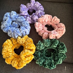 Puede incluir: Cinco coleteros de felpa en varios colores: azul, morado, rosa, amarillo y verde. Estos suaves accesorios para el cabello con textura están hechos de un material aterciopelado. Los coleteros están colocados sobre una superficie gris.