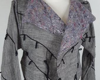 Peut inclure: Une veste courte grise et noire avec un design unique comprenant des bretelles noires et un col en tissu gris. La veste a une coupe ample et une ourlet légèrement asymétrique.