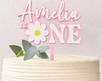 May include: A white frosted cake on a white pedestal plate, topped with a pink and white cake topper that reads "Amelia is One". The topper features a daisy and greenery. A perfect cake for a first birthday celebration.