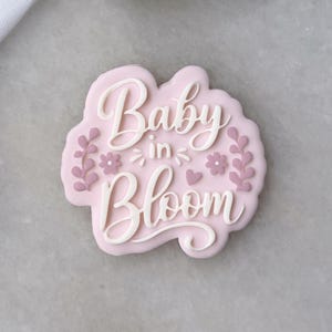 Może przedstawiać: Jasnoróżowa foremka do ciastek z napisem "Baby in Bloom" w białym skrypcie. Projekt zawiera kwiatowe i liściaste akcenty. Foremka znajduje się na jasnoszarej powierzchni z zielonymi liśćmi w tle.