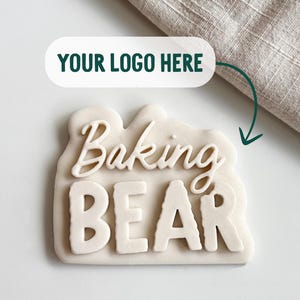 May include: A white cookie cutter or stamp featuring the embossed text Baking BEAR in a combination of script and block lettering. Above the object, a white rectangular label with rounded corners displays the text YOUR LOGO HERE.