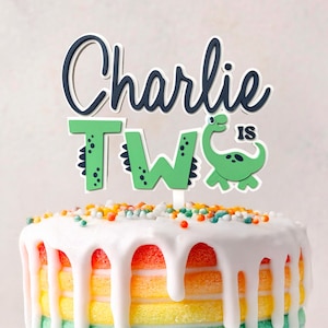 Peut inclure: Un gâteau d'anniversaire coloré avec un glaçage blanc dégoulinant. Le gâteau a des couches arc-en-ciel et est surmonté de vermicelles et d'un cake topper avec l'inscription "Charlie a deux ans" et un dinosaure.