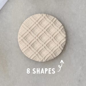May include: A beige, circular cookie cutter with a plaid pattern, next to a rectangular cutter and a round cutter. The image includes the text "8 SHAPES". Green leaves and a white cloth are in the background.
