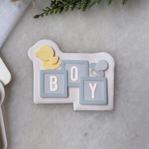 Può includere: Un biscotto di colore chiaro decorato con la parola "BOY" scritta con blocchi blu e un'anatra gialla. Il biscotto è decorato con accenti blu chiaro e giallo. Formine per biscotti sono visibili nell'immagine.