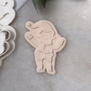 May include: A light beige cookie cutter in the shape of a cartoon elf with arms raised. The elf wears a hat, belt, and boots. Another cookie cutter is visible to the left. The background is a light-colored surface with greenery.