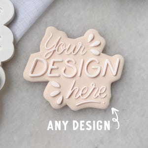 May include: A light beige custom cookie cutter featuring the embossed text "Your DESIGN here" in a script and sans-serif font combination. The cutter has an irregular, cloud-like shape with small decorative leaf-like accents. Below, the text "ANY DESIGN" is visible.