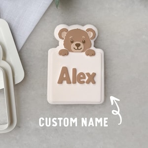 Puede incluir: Un cortador de galletas de color claro y una galleta con forma de oso con el nombre "Alex" en letras marrones. El oso es marrón y asoma por encima de un letrero rectangular. Las palabras "CUSTOM NAME" están debajo.