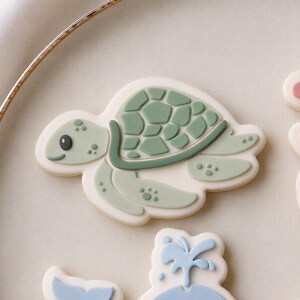 May include: A sea turtle biscuit decorated with sage green icing, featuring a detailed shell pattern and a small black eye. The biscuit is shaped like a swimming turtle with flippers and a smooth off-white base, presented on a round ceramic plate.