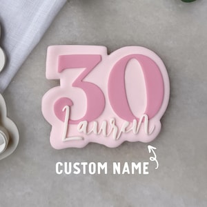 May include: A light pink cookie cutter with the number 30 in a darker pink, and the name "Lauren" in white script. The text "CUSTOM NAME" is below the cookie. The cookie cutter is for making custom cookies.