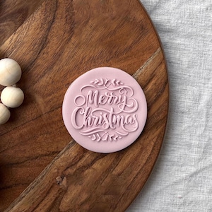 Plastic cookie cutter and fondant stamp with a custom design saying "Merry Christmas", made of durable plastic and shown on a countertop.