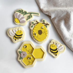 May include: A collection of bee-themed cookies. The cookies feature a yellow honeycomb, a beehive, and smiling bees with black and yellow stripes. Some cookies have floral accents. The cookies are on a white surface.