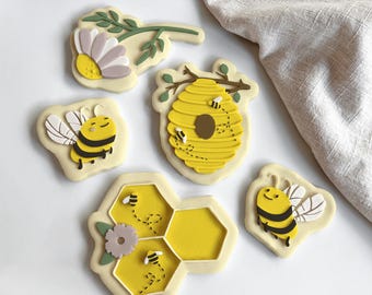Può includere: Una collezione di biscotti a tema api. I biscotti presentano un favo giallo, un alveare e api sorridenti con strisce nere e gialle. Alcuni biscotti hanno accenti floreali. I biscotti sono su una superficie bianca.