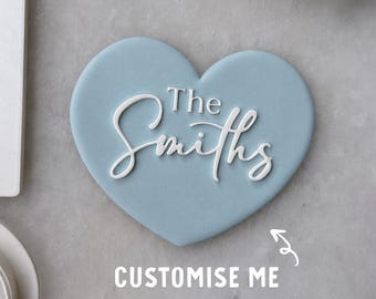 May include: A light blue heart-shaped plaque featuring the white raised text "The Smiths" in a combination of serif and script fonts. The plaque is smooth and matte, with a clean, simple design that highlights the personalized lettering.