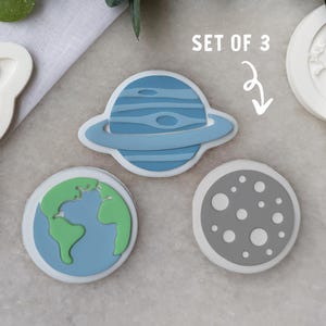 May include: A set of three space-themed cookie cutters. The set includes a blue planet with rings, a green and blue Earth, and a gray moon with white craters. The text "SET OF 3" is visible.
