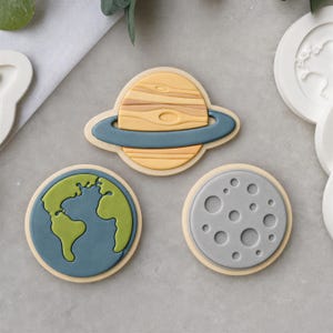 May include: Three circular cookie cutters featuring planetary designs. One cutter depicts a yellow and tan ringed planet, another shows a blue and green Earth, and the third displays a gray moon with circular craters on a light background.