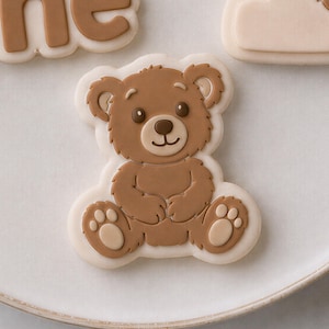 K&ouml;nnte beinhalten: Ein b&auml;renf&ouml;rmiger Keks, verziert mit brauner und cremefarbener Glasur. Der B&auml;r hat ein l&auml;chelndes Gesicht, dunkelbraune Augen und eine hellbraune Schnauze. Die Pfoten haben hellbraune Ballen. Der Keks ist mit cremefarbener Glasur umrandet.