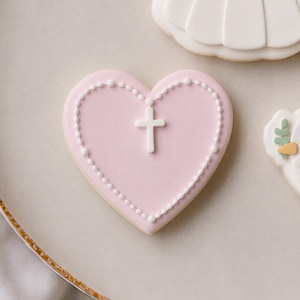 May include: A heart-shaped cookie with a smooth light pink icing base. The cookie features a white piped beaded border that follows the heart's edge and a central white cross emblem made of icing, creating a clean and symmetrical design.
