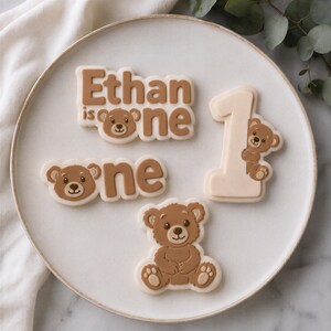 K&ouml;nnte beinhalten: Vier verzierte Kekse auf einem Teller. Ein Keks tr&auml;gt die Aufschrift Ethan is one mit einem B&auml;rengesicht. Ein weiterer zeigt one mit einem B&auml;rengesicht. Ein dritter ist als Zahl 1 mit einem B&auml;ren geformt. Der vierte ist ein B&auml;renkeks.