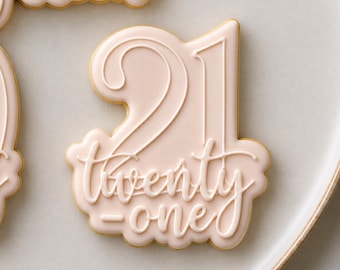 May include: A light pink sugar cookie shaped like the number 21, featuring raised white icing in a script font that spells out twenty-one. The cookie has a smooth, matte finish and a scalloped edge, with a portion of another cookie visible.
