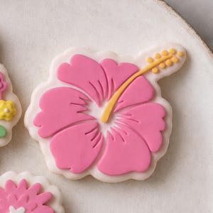 May include: A sugar cookie shaped like a hibiscus flower with bright pink icing petals and a yellow stamen extending from the center. The flower design is layered over a white icing base, creating a distinct floral shape with smooth edges.