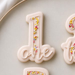 Puede incluir: Una galleta de color beige claro con forma de número uno, decorada con glaseado blanco y la palabra one en letra cursiva. El centro del número está relleno de pequeñas chispas redondas y coloridas en tonos amarillos, rosas y azules.