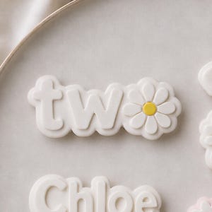 Pode incluir: Um biscoito branco com um design em relevo apresentando as letras minúsculas 'two' seguidas por uma forma de margarida com um centro amarelo brilhante. O biscoito tem um acabamento fosco liso e uma borda arredondada e festonada.