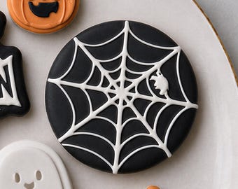 May include: A round cookie decorated with black icing and a white piped spiderweb design. The web features a small white spider detail. Other Halloween-themed cookies, including a pumpkin and a ghost, are partially visible in the frame.