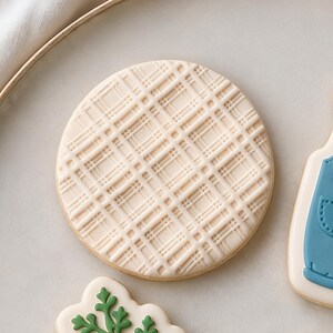May include: A round sugar cookie decorated with a raised, off-white icing pattern featuring a grid of intersecting lines that create a textured plaid design across the entire surface of the circular biscuit.