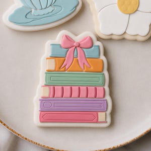 以下が含まれることがあります： 本のような形をした5枚の長方形のクッキーが積み重なっており、水色、オレンジ、セージグリーン、薄いピンク、ラベンダー色の滑らかなアイシングで装飾されています。積み重ねられたクッキーの上部には大きな薄いピンクのアイシングのリボンがあり、皿の中央に置かれています。