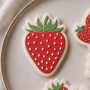 Peut inclure: Un biscuit en forme de fraise avec un glaçage rouge lisse, des points de glaçage blancs représentant les graines et des feuilles en glaçage vert sur le dessus. Le biscuit a une bordure festonnée crème sur une assiette claire à bord doré.