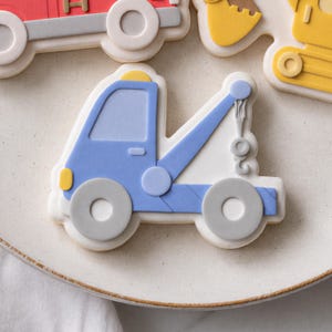 May include: A sugar cookie shaped like a blue tow truck with a yellow light on top, grey wheels, and a white base. The truck features a blue crane arm with a hook, and the design is rendered in smooth icing with clean, defined edges and details.