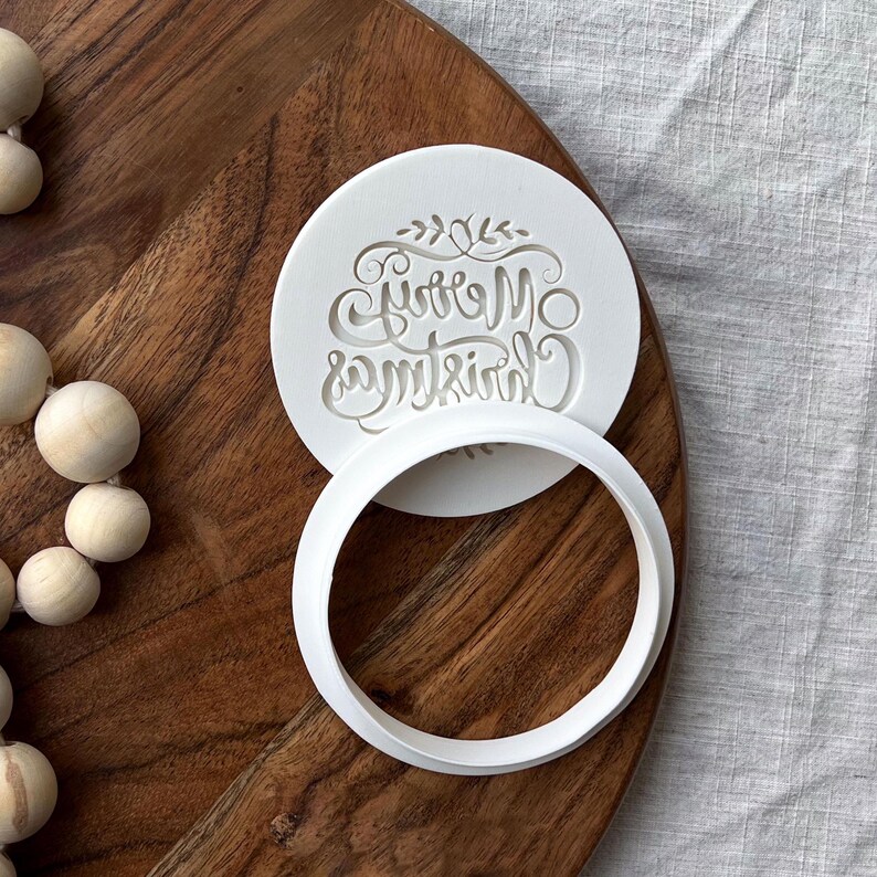 Plastic cookie cutter and fondant stamp with a custom design saying "Merry Christmas", made of durable plastic and shown on a countertop.