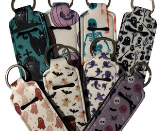May include: A collection of Halloween-themed lip balm holders with key rings. The holders feature various patterns, including ghosts, bats, skeletons, and pumpkins. Colors include teal, purple, white, and pink. The Wildflower Avenue logo is at the bottom.