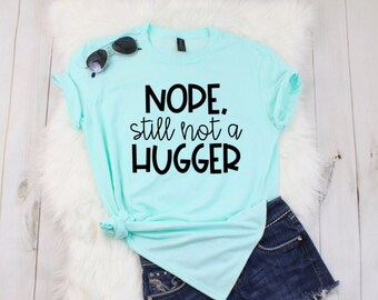 May include: A light blue t-shirt with the text "NOPE, still not a HUGGER" in black lettering. The shirt is laid out on a white fuzzy surface with a pair of sunglasses and denim shorts.