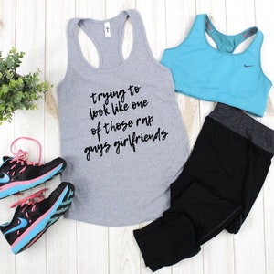 May include: Gray tank top with the text "trying to look like one of those rap guys girlfriends".  Black leggings, a blue sports bra, and black and pink running shoes are also pictured.